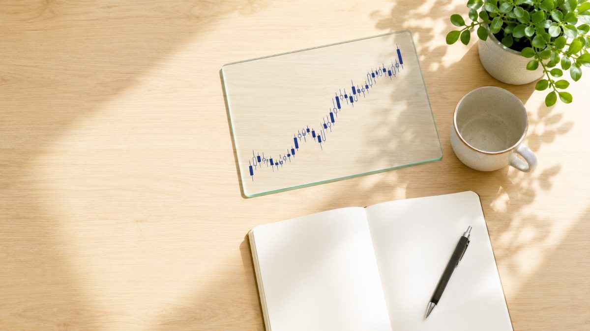FTO（Forex Tester Online）で過去検証をする朝の机を真上から見たイメージ。上昇チャートを映したガラスのパネルとマグカップ、開いたノート